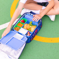 A child sits on a green outdoor court floor next to an open Montii Large Insulated lunch bag filled with a bento box, sandwich, fruit, and snacks. 