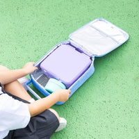 A child sits beside an open Montii Darling Bows Large Insulated Lunch Bag on a green textured surface, holding a purple lunchbox and a 200ml Montii Food Jar inside the bag.
