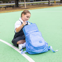 A young girl in a school uniform sits on a green outdoor court putting a Montii Darling Bows Large Insulated Lunch Bag into her backpack.