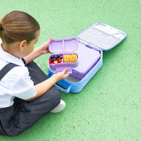 A child in a school uniform sits on a green textured surface next to an open Montii Darling Bows Large Insulated Lunch Bag, opening a Lilac Snack Box filled with fruit and crackers.