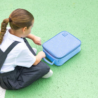 A child in a white shirt and dark overalls sits on a green textured surface next to a Montii Darling Bows Large Insulated Lunch Bag