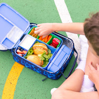 A child's open Montii High Voltage Large Insulated Lunch Bag filled with a packed school lunch sits on a green outdoor court. The child's hand is reaching into the Montii Lunchbox, which contain a burger, carrots, strawberries, and other snacks.