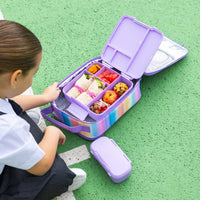 A child sits beside an openMontii Sunset Skies Large Insulated Lunch Bag on a green textured surface. There is an open lunch box filled with wraps, fruit, and snacks.