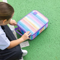 A child wearing a white shirt and dark pants sits on a green textured surface next to her Montii Sunset Skies Large Insulated Lunch Bag