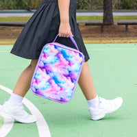 A person in a black pleated skirt and white sneakers carrying the Montii Large lunch bag with a pastel pink, purple, and blue cloud pattern on a green outdoor court.