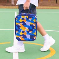 A child carrying a Montii Camo Trail Insulated lunch bag on a green outdoor court.  The Montii lunch bag includes an ice pack