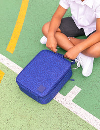 A child in a white school uniform sits on a green sports court, opening a Montii Shapeshifter Insulated lunch bag.