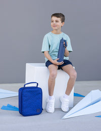 A young boy sits on a white block holding a Montii water bottle, with a Montii Shapeshifter Insulated lunch bag on the floor next to him. The scene is set against a neutral gray background with paper airplanes on the floor.