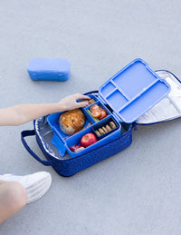 The Montii Large Insulated Lunchbag on a gray concrete surface, shown open with a child's hand placing food inside the lunchbox. 