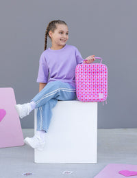 A young girl with braided hair sits on a white block wearing a lavender t-shirt and light blue jeans with white stripes. She is holding a Queen of Hearts Montii Co Insulated Lunchbag.