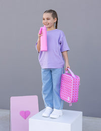 A young girl stands on a white pedestal against a gray studio background, holding a pink water bottle and a Queen of Hearts Montii lunch bag. She is wearing a lavender t-shirt, light blue jeans, and white sneakers.