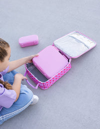 A child sits on a gray concrete surface putting a Montii Bento Lunchbox into the Queen of Hearts Insulated Lunch bag. A matching Montii Snack box is placed nearby.