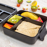 Montii Feast lunchbox with compartments containing a sandwich, fruit, and vegetables on a light surface.