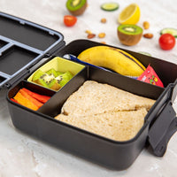 Montii Feast lunchbox with compartments containing a sandwich, fruits, and vegetables on a light surface.