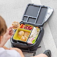 Person opening a Montii Feast lunch box with various food items on a light gray surface.
