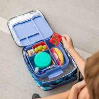Montii Feast bento lunch box with compartments filled with snacks and a hand holding a snack card on a gray surface.