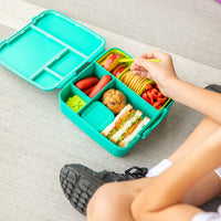 Person opening a Montii Feast lunchbox filled with food on a light gray surface.