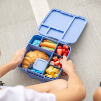 Montii Feast Bento Lunchbox with compartments filled with food, held by a child's hands on a light gray surface.