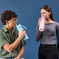 Two people drinking from the Lagoon BBox Drink Bottle and the Pink BBox Drink bottle against a blue background