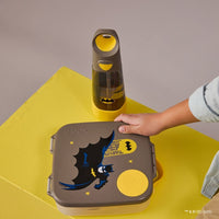 BBox Batman-themed lunch box and bottle on a yellow surface with a child's hand reaching for the lunch box.