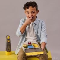 Child holding a BBox Batman themed lunchbox with snacks, sitting on a yellow stool against a gray background