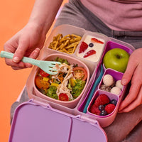 Child using a green fork to eat from a BBox Sugar Plum bento lunchbox filled with food on an orange background