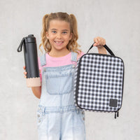 Young girl holding a Montii black water bottle and Montii Black Gingham lunch bag against a light gray background