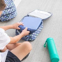 Child packing a Montii lunch box into a Montii Checkmate Insulated Lunchbag on a gray floor.