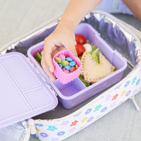 MontiiCo Lilac Lunchbox with compartments filled with snacks, including a hand reaching in to grab a container of colorful candies.  All packed inside a Montii Daisy Dream Large Insulated Lunchbag