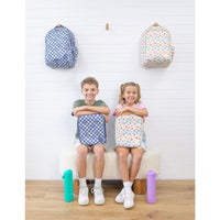 Two children sitting on a bench with the Montii Large Insulated Lunchbags and the Junior backpacks on a white wall background