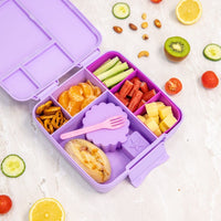 Montii Lilac bento box with compartments filled with snacks and a pink fork, surrounded by fresh fruits and vegetables on a white surface.