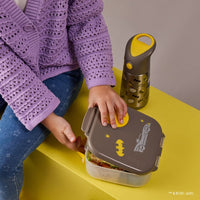 Child opening a BBox lunchbox with a Batman design on a yellow surface, next to a BBox Batman-themed water bottle.
